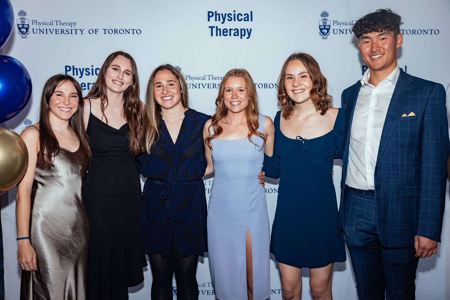 Group of six MScPT graduates standing together in front of a photo wall.